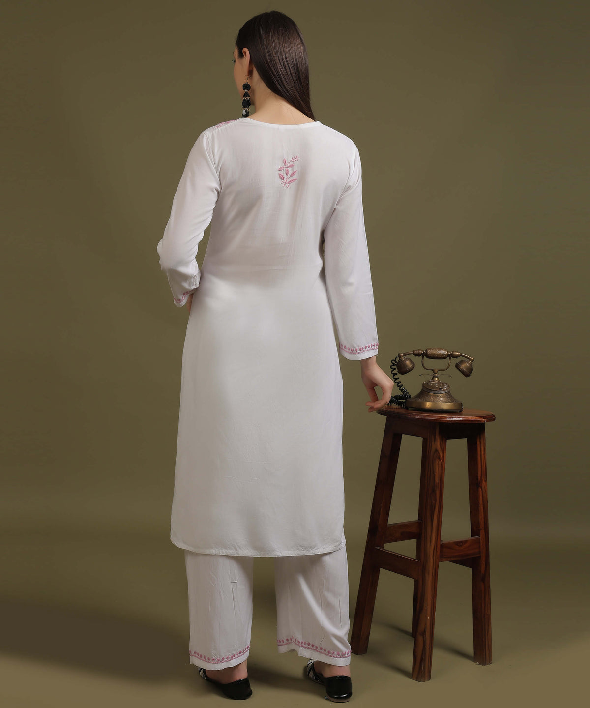 Back view of model in NOORI white rayon chikankari kurta with pink back motif next to vintage telephone on wooden stool by Noor-e-Awadh